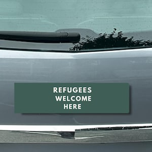 Peut inclure: Un autocollant rectangulaire vert fonc&eacute; avec le texte blanc &laquo;&nbsp;REFUGEES WELCOME HERE&nbsp;&raquo;. L'autocollant est appos&eacute; sur le devant d'une voiture grise, sous le pare-brise.