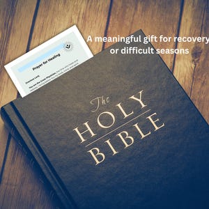 Puede incluir: Una Biblia azul oscuro con letras doradas, titulada "The Holy Bible". Se inserta un marcador con el texto "Prayer for Healing". La imagen incluye el texto "Un regalo significativo para la recuperaci&oacute;n o las estaciones dif&iacute;ciles."