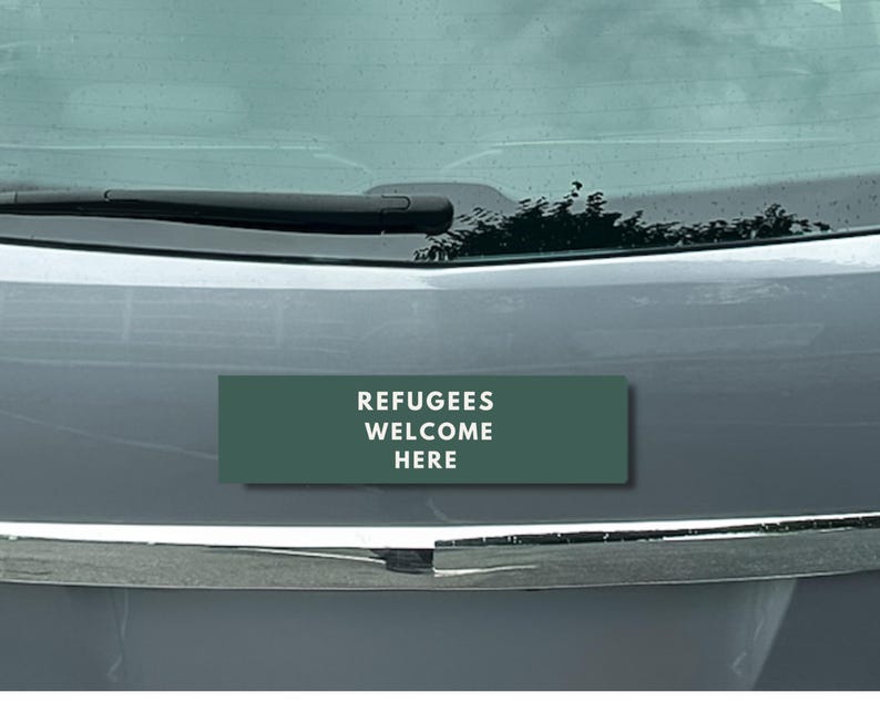 Peut inclure: Un autocollant rectangulaire vert fonc&eacute; avec le texte blanc &laquo;&nbsp;REFUGEES WELCOME HERE&nbsp;&raquo;. L'autocollant est appos&eacute; sur le devant d'une voiture grise, sous le pare-brise.