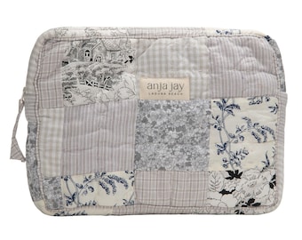 May include: A quilted, patchwork cosmetic bag in shades of gray and white. The bag features various fabric patterns, including floral, striped, and toile designs. The label reads "anja jay laguna beach".
