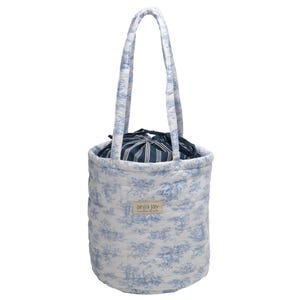 May include: A quilted tote bag with a blue and white toile pattern. The bag has a cylindrical shape with a drawstring closure and a striped interior. The handles are also quilted and patterned. The brand name "anja jay" is printed on the front.