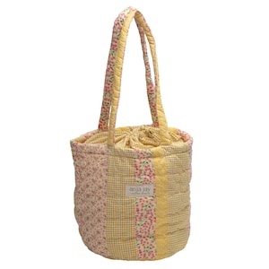 May include: A quilted tote bag with a floral and gingham pattern in yellow, pink, and cream. The bag has two handles and a drawstring closure. The label reads "anja jay LONDON BEACH".
