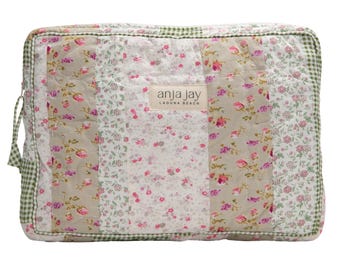 May include: A rectangular quilted bag with a patchwork design. The bag features floral patterns in pink and white, with beige and green accents. The edges are trimmed with a green gingham pattern. The label reads "anja jay LAGUNA BEACH".
