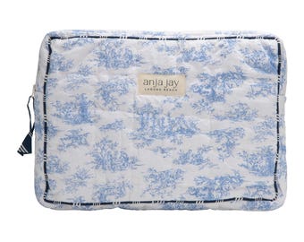 May include: A rectangular, quilted cosmetic bag with a blue and white toile pattern. The bag features a navy blue zipper pull and trim. The label "anja jay laguna beach" is centered on the front of the bag.