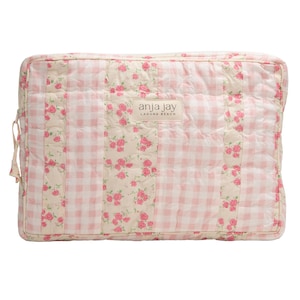 May include: A rectangular quilted bag with a floral and gingham pattern. The bag features a cream-colored background with pink roses and a pink and white gingham pattern. The brand name "anja jay LAGUNA BEACH" is displayed on a rectangular label.