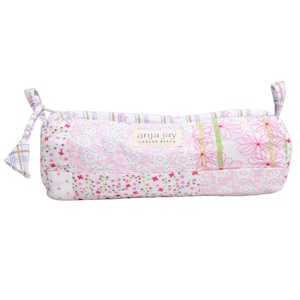 May include: A pink and white quilted pencil case with floral and geometric patterns. The case has a rectangular shape with rounded ends and a drawstring closure. The brand name "anja jay laguna beach" is printed on the front.