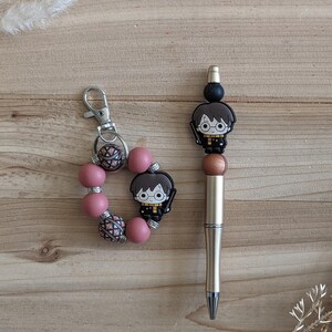 Peut inclure: Un stylo doré et un porte-clés à perles roses sur le thème d'Harry Potter. Le stylo a un dessus noir et doré avec un charme de personnage d'Harry Potter. Le porte-clés comporte des perles roses et un charme d'Harry Potter. Le fond est en bois brun clair.