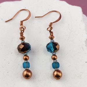 Può includere: Un paio di orecchini pendenti con monachelle color rame, una perla di cristallo blu scuro sfaccettata, una piccola perla in rame metallizzato, una perla azzurra opaca e una sfera in rame liscio alla base di ogni orecchino.