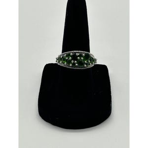 May include: A silver ring with a row of round, vibrant green gemstones. The ring is displayed on a black jewelry stand, showcasing its design and color. The gemstones are set closely together, creating a continuous band of green.