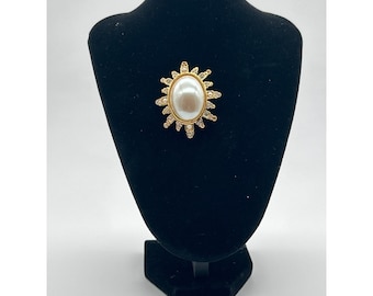 May include: A gold-toned brooch featuring a large, oval faux pearl center. The pearl is surrounded by a sunburst design with small, clear crystals and gold-colored metal accents. The brooch is displayed on a black mannequin.