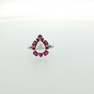 May include: A silver-toned ring featuring a central pear-shaped iridescent white stone surrounded by a halo of small, oval-cut vibrant pink stones. The metal band is split at the shoulders, leading to a polished finish on the ring's setting.