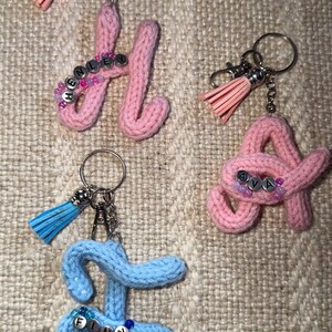 Può includere: Portachiavi fatti a mano in filato rosa e blu, ciascuno a forma di lettera. I portachiavi rosa compongono "HENLEY" e "AVA", mentre il portachiavi blu compone "FINN". Ogni portachiavi ha un nappa e piccole perline.