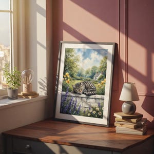 May include: Framed artwork featuring a tabby cat resting on a stone wall in a garden scene. The painting depicts a white cottage, yellow flowers, and a blue sky. The artwork is displayed on a wooden table near a window.