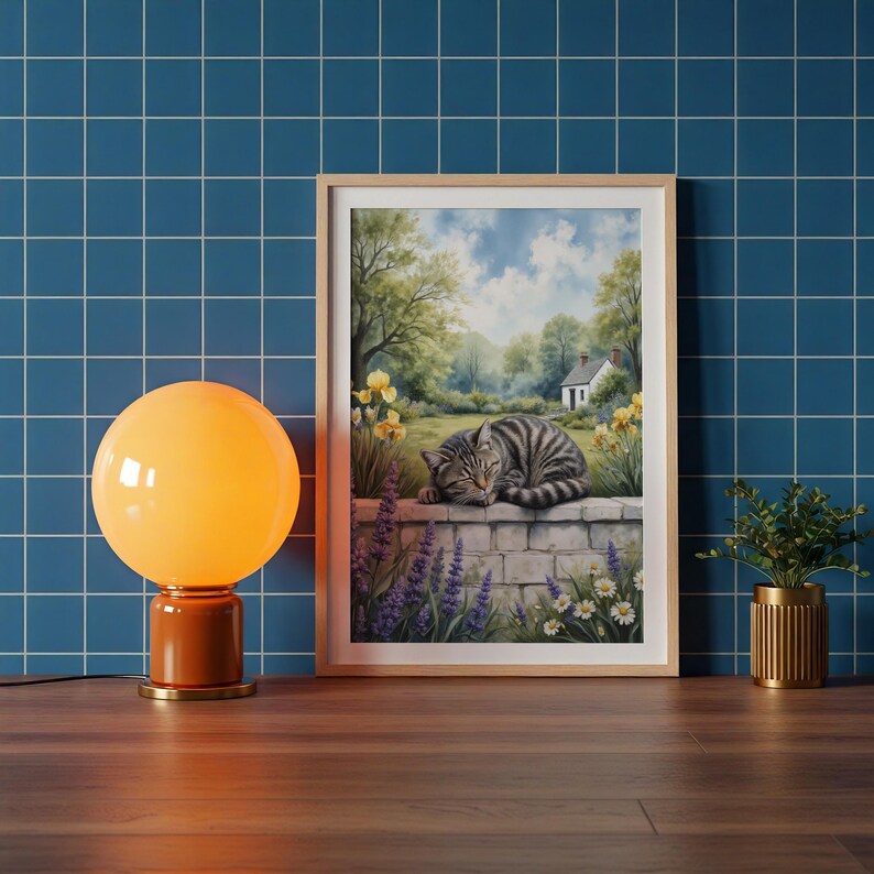 May include: A framed print of a tabby cat sleeping on a stone wall in a garden. The artwork features a serene landscape with a white house, trees, and flowers. An orange globe lamp and a small potted plant are on a wooden surface.