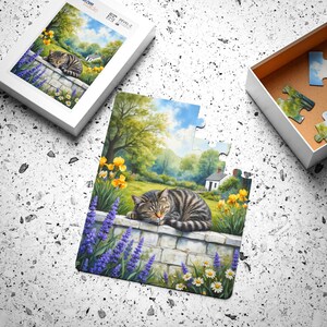 May include: A jigsaw puzzle featuring a tabby cat resting on a stone wall, surrounded by purple lavender, yellow flowers, and a scenic landscape. The puzzle is partially assembled, with a box containing the remaining pieces.