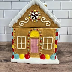 Könnte beinhalten: Ein Keramik-Lebkuchenhaus-Keksdose mit weißem, schneebedecktem Dach und dekorativen Zuckergussdetails. Das Haus hat eine rosa Tür, Fenster und bunte Süßigkeitenakzente. Das Glas ist so gestaltet, dass es wie ein festlicher Leckerbissen aussieht.
