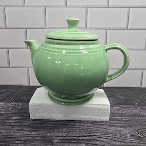 May include: A light green ceramic teapot with a rounded body, a matching lid with a small knob handle, a curved spout, and a side handle. The teapot features concentric ring details around the lid and the upper portion of the main body.