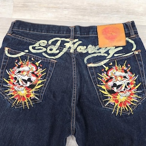 May include: Dark blue denim jeans featuring a tan leather patch on the back waistband. The back pockets display colorful embroidered skull graphics with yellow and red sunburst patterns, and the Ed Hardy logo is stitched in light green.