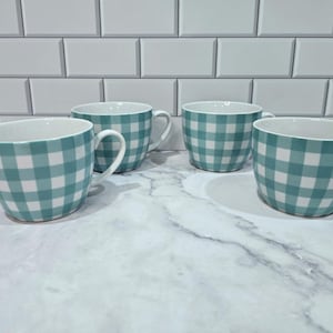 May include: Four ceramic mugs with a teal and white gingham pattern. The mugs have a rounded shape with a white interior and a curved handle. They are arranged on a marble-like surface in front of a white tile wall.