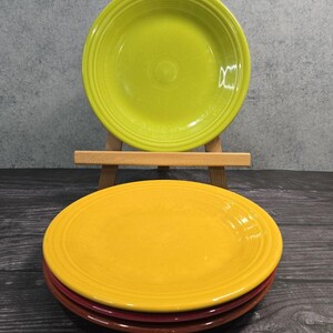May include: A stack of colorful ceramic plates in shades of yellow, green, and red. The plates have a classic design with a slightly raised rim. One green plate is displayed on a small wooden easel. The plates are arranged on a dark wooden surface.