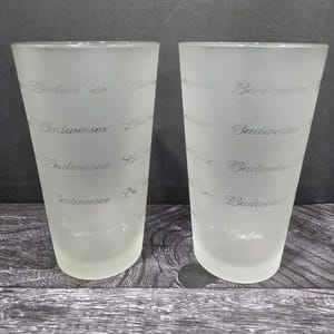 May include: Two frosted glass Budweiser beer glasses. The glasses have a tapered shape and the Budweiser logo is repeated in a pattern on the glass. The glasses are on a dark wood surface.