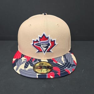 Può includere: Cappellino da baseball beige con visiera mimetica rossa, grigia e blu navy. Il cappellino presenta un logo della squadra con un uccello blu e una foglia d'acero rossa. Taglia 7 1/4.