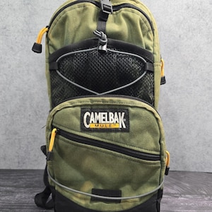 以下が含まれることがあります： オリーブグリーンのCamelBak M.U.L.E.ハイドレーションバックパック。黒いメッシュのフロントポケット、CamelBak M.U.L.E.のロゴが入った黒いジッパー付きコンパートメント、黄色のジッパープル、上部の黒いバックル、収納用のグレーの伸縮性コードシステムが特徴です。