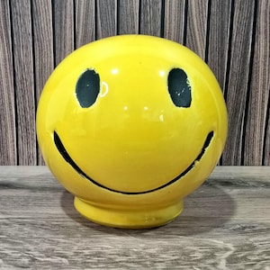 Puede incluir: Una figura de cerámica amarilla con una cara sonriente con ojos negros y una boca negra curvada. La cara sonriente es redonda y se asienta sobre una pequeña base circular. Fondo de madera.