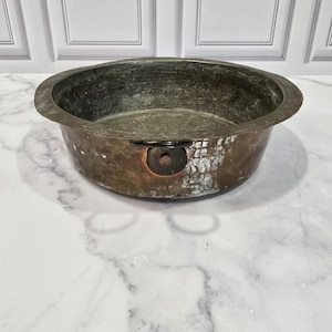May include: An antique copper bowl with a wide rim and a dark, aged patina. The bowl features a small, riveted handle and shows signs of wear. The interior has a greenish hue, and the exterior is a mix of copper and verdigris, indicating age and use.