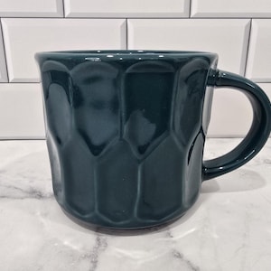 May include: A teal ceramic mug with a textured, geometric pattern. The mug has a rounded handle and a glossy finish. The mug is sitting on a white surface in front of a white tiled wall.