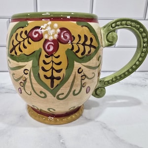 Puede incluir: Una taza de cerámica decorativa con cuerpo redondeado y asa verde. La taza presenta una base beige con diseños florales amarillos y verdes, acentuados con detalles rojos y morados. El borde y la base están adornados en verde y dorado.