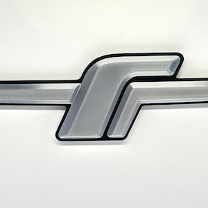 May include: A silver-colored emblem featuring a stylized letter F design with a horizontal bar extending from the center. The emblem has a smooth, metallic finish with a distinct black border outlining the entire shape against a plain background.