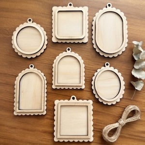 May include: A collection of seven light-colored wooden frames with scalloped edges in various shapes including square, round, oval, and arched designs. Each frame features a central cutout and a small hole at the top for hanging, accompanied by twine.