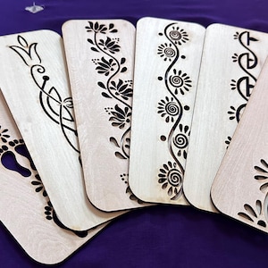 May include: A collection of six wooden bookmarks with intricate laser-cut designs. The designs include floral patterns, swirls, and footprints. The bookmarks are light-colored wood and are arranged on a purple surface.
