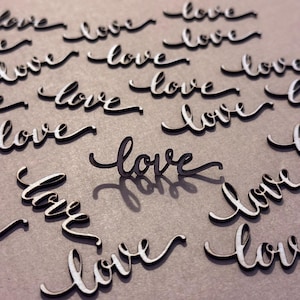May include: A collection of wooden cutouts spelling the word "love" in a cursive font. The letters are white with a dark edge, scattered on a neutral surface, creating a romantic theme.