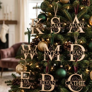 May include: Wooden Christmas tree ornaments with names like Adam, Diane, and Cathie. The ornaments are in the shape of letters with decorative swirls and are hanging on a decorated Christmas tree with green and gold ornaments.