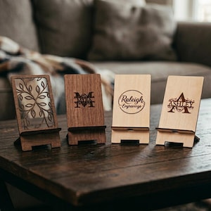 May include: Four rectangular wooden phone stands arranged in a row. The first is dark wood with a laser-cut tree design. The second is medium-toned wood with the name Myra. The third is light wood with the text Raleigh Engravings. The fourth is light wood with the name Ashley.