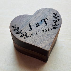 Puede incluir: Caja de madera en forma de corazón con las iniciales grabadas "J & T" y la fecha "10.17.2025". La caja tiene un acabado de madera oscura con una parte superior gris más clara y un diseño de hojas decorativas. Un recuerdo único.