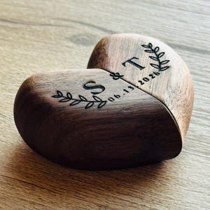 Puede incluir: Una caja de madera en forma de corazón, dividida en dos, con iniciales y una fecha grabadas. La caja es de madera oscura y presenta las iniciales "S" y "T" con la fecha "06.13.2026" y detalles de hojas decorativas.
