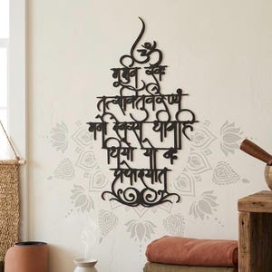 May include: Black metal wall art with intricate script, displayed on a pale wall with a delicate floral design. The script is a stylized mantra, ideal for meditation areas or spiritual decoration.