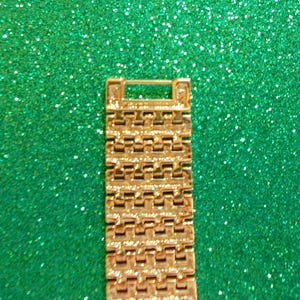 May include: A gold-colored watch band with a rectangular design. The band is made of multiple small, connected rectangular links. The background is a sparkly, glittery green.