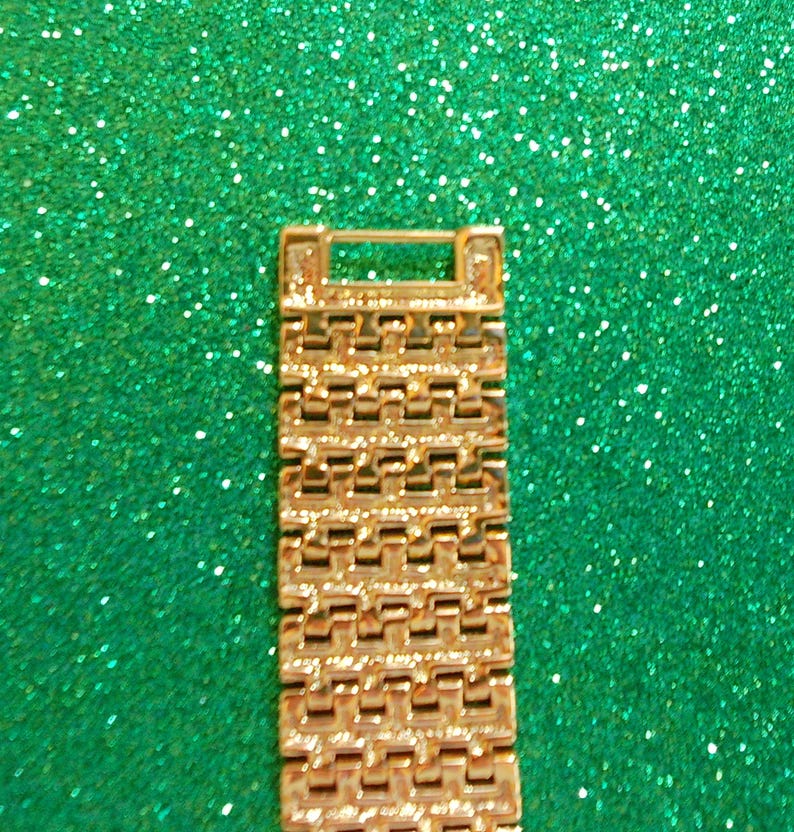 May include: A gold-colored watch band with a rectangular design. The band is made of multiple small, connected rectangular links. The background is a sparkly, glittery green.