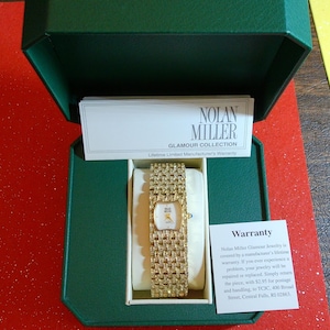 May include: A gold-toned Nolan Miller Glamour Collection watch with a rectangular face and a wide, textured band, presented in a green box. The watch is displayed on a cream-colored cushion, with a warranty card visible.