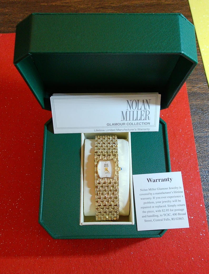 May include: A gold-toned Nolan Miller Glamour Collection watch with a rectangular face and a wide, textured band, presented in a green box. The watch is displayed on a cream-colored cushion, with a warranty card visible.