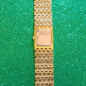 May include: Gold-toned rectangular watch with a matching link band. The watch face is a light color, and the band has a repeating pattern. The watch is displayed against a green, glittery background.