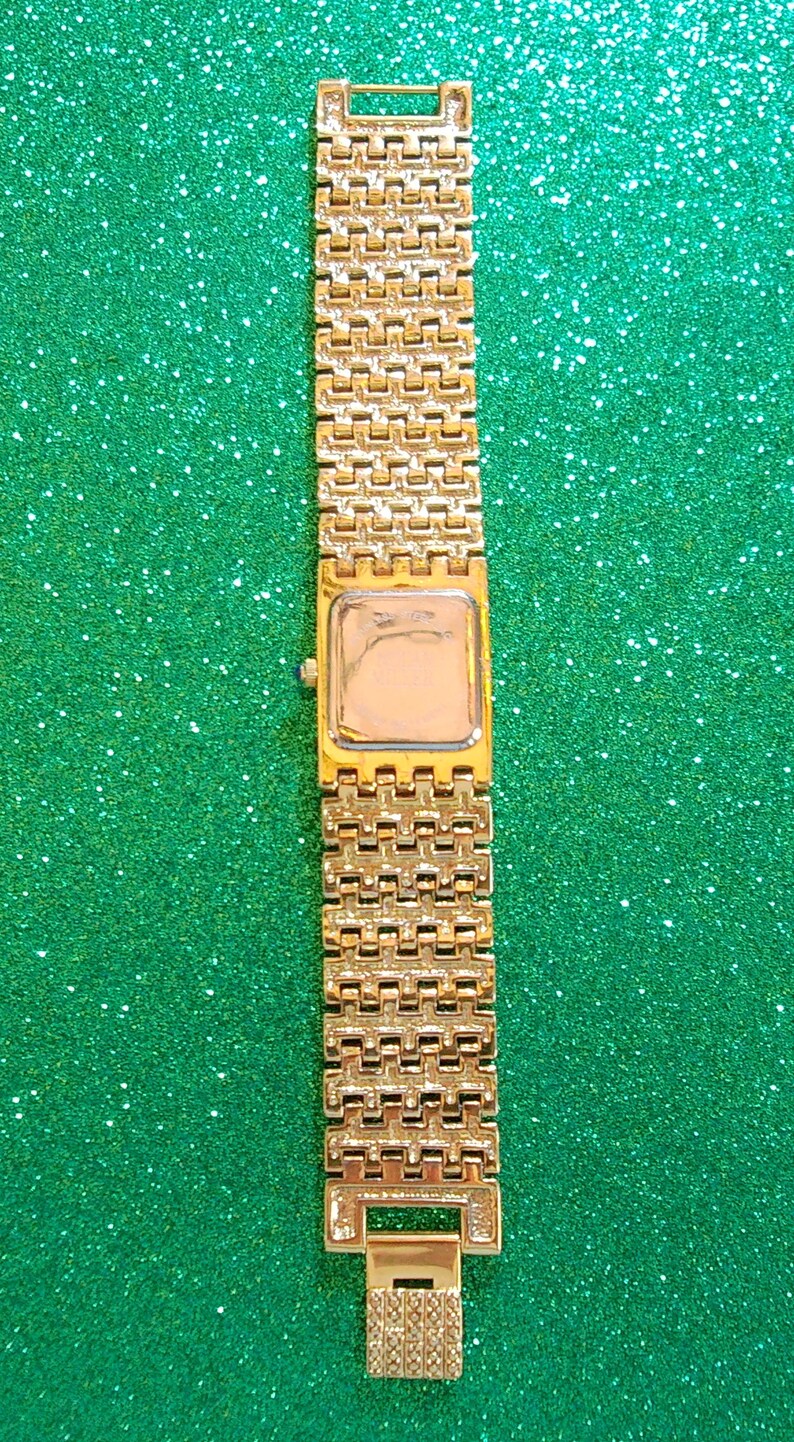 May include: Gold-toned rectangular watch with a matching link band. The watch face is a light color, and the band has a repeating pattern. The watch is displayed against a green, glittery background.