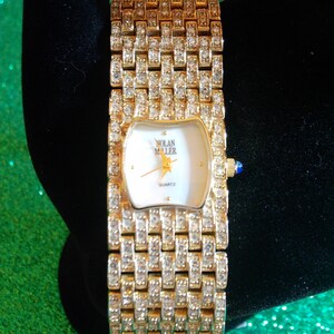 May include: A gold-toned Nolan Miller quartz watch with a rectangular face and a band encrusted with clear crystals. The watch face is white with gold hands and a star emblem. A small blue gem accents the side.