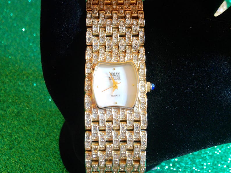 May include: A gold-toned Nolan Miller quartz watch with a rectangular face and a band encrusted with clear crystals. The watch face is white with gold hands and a star emblem. A small blue gem accents the side.