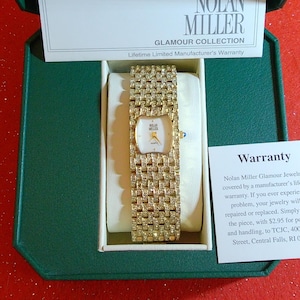 May include: A gold-toned Nolan Miller Glamour Collection watch with a rectangular face and a band encrusted with sparkling stones. The watch is presented in a green box with a white interior, accompanied by a warranty card. The watch face has a white background with gold hands.