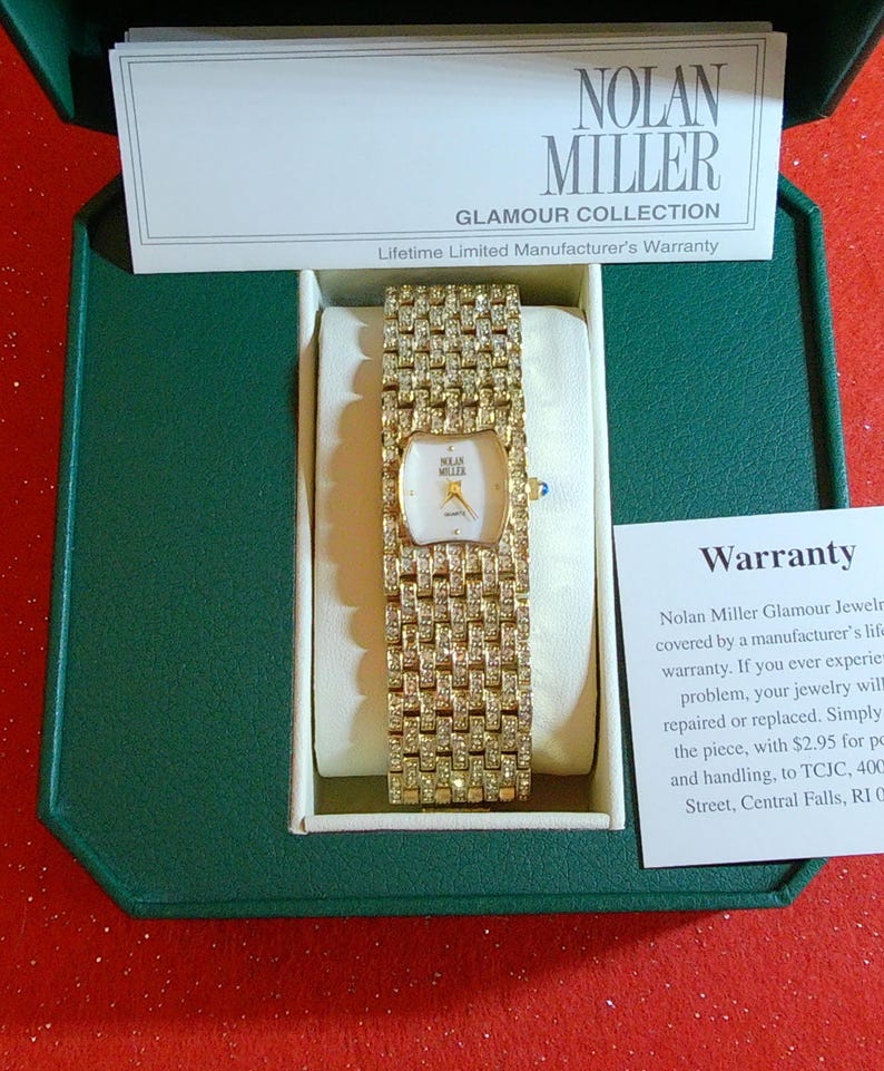 May include: A gold-toned Nolan Miller Glamour Collection watch with a rectangular face and a band encrusted with sparkling stones. The watch is presented in a green box with a white interior, accompanied by a warranty card. The watch face has a white background with gold hands.